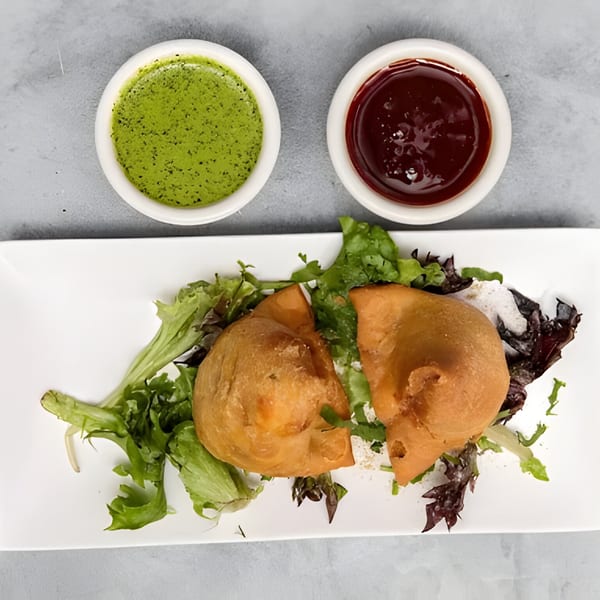 Two golden-brown samosas on a bed of fresh greens, served with green chutney and tamarind sauce.
